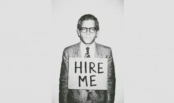 A man in a suit and glasses wearing a sign that says "hire me" 